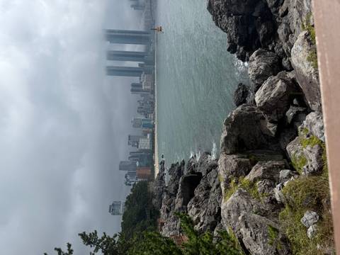       Rocky coastline with rough waves and a misty modern city skyline of tall skyscrapers in the background.
  