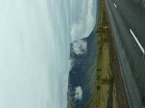       Sheer basalt cliff shrouded in low clouds rising from a grassy plain beside a wet roadway
  