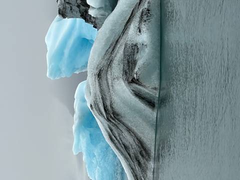       Vertical close-up of a blue and white iceberg with sharp ridges floating in calm grey water
  