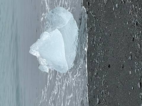       Clear block of glacial ice washed up on black volcanic sand as gentle waves lap the shore
  