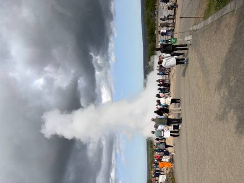       Crowds watch a powerful white geyser erupting against a backdrop of dramatic dark clouds
  