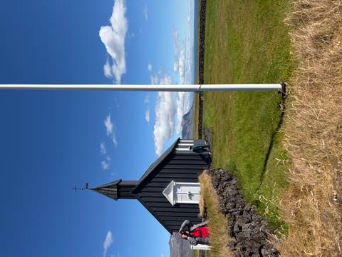       Black wooden church at Búðir with a lone visitor nearby, bright blue sky and distant mountain ridge
  