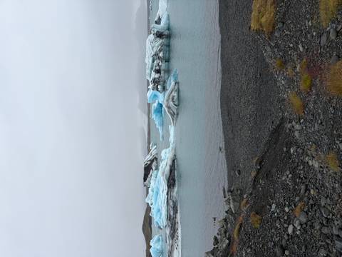       Wide glacier lagoon scene with blue icebergs floating in calm teal water bordered by dark pebble shore
  