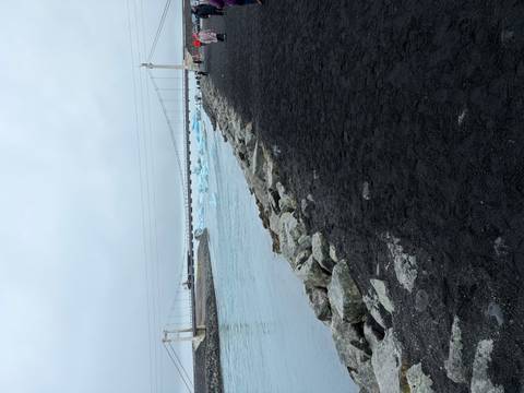       Suspension bridge over a glacial outflow channel with black sand shoreline and distant icebergs
  