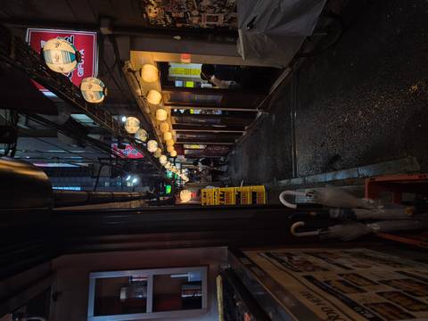       Narrow lantern-lit alley lined with small eateries and bars on a rainy night.
  