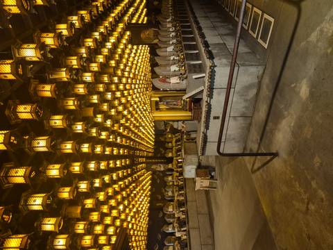       Hall filled with hundreds of glowing golden lanterns and stone statues in soft light.
  