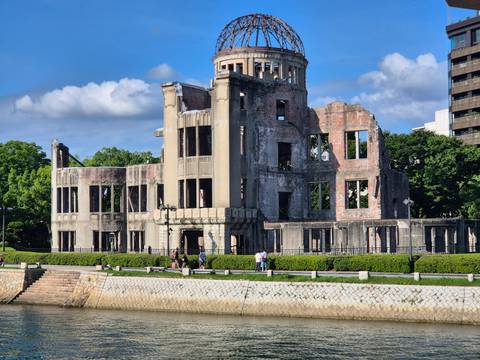       Historic A-Bomb Dome ruins stand beside a river under bright blue skies.
  