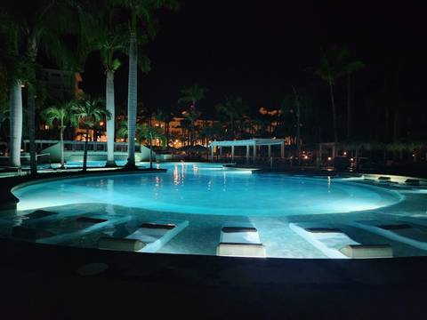       Resort swimming pool glows turquoise under night lights surrounded by palms and cabanas.
  