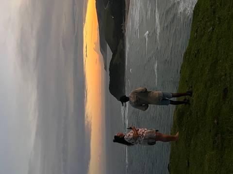       Two travelers standing on grassy cliff watching a colorful sunset over rugged coastline.
  
