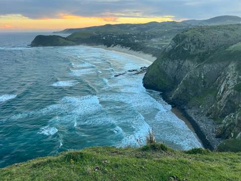       Aerial coastal view of rolling surf hitting a long sandy beach backed by green hills.
  