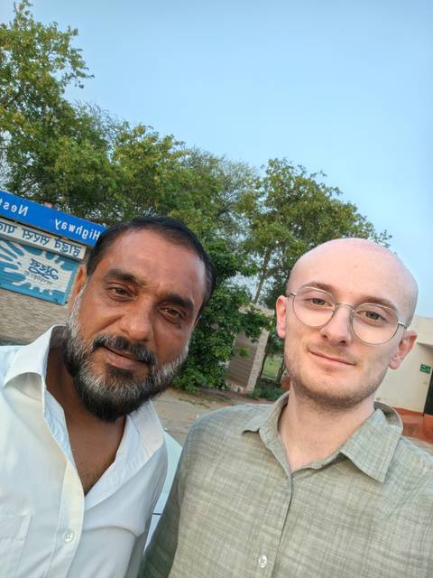       Close-up selfie of two men smiling outdoors with trees and a signboard behind them.
  