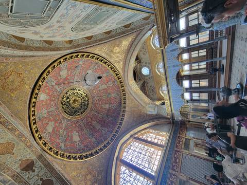       Lavish Ottoman hall with decorative gilded ceiling and visitors observing the artwork.
  