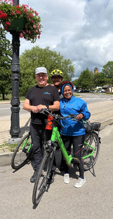       Cyclists pose with green e-bikes on a roadside under sunny skies.
  