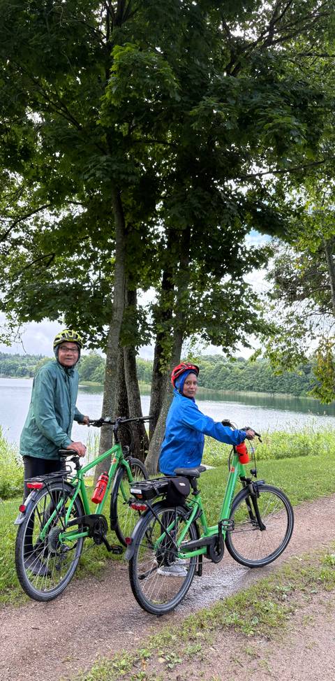       Cycling pair stop by a lakeside path shaded by tall trees on a cloudy day.
  
