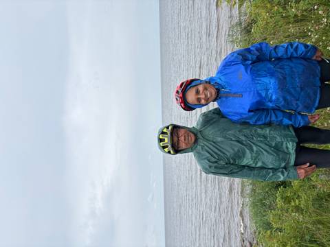       Couple in rain gear and helmets stands at a windy Baltic seashore under grey skies.
  