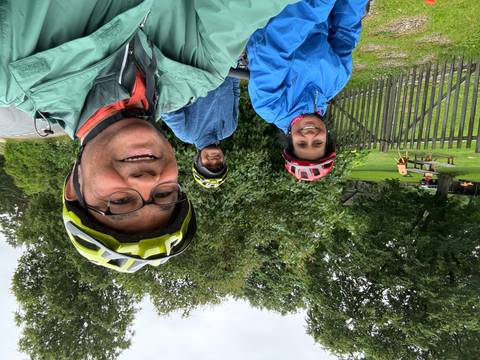       Cyclists smile for a selfie beside lush greenery after a ride on a rainy day.
  