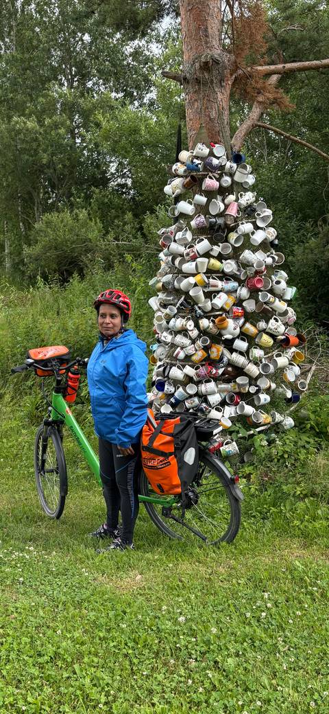       Cyclist in a blue rain jacket and red helmet stands with a green touring bike beside a sculptural tower of stacked coffee mugs in a lush, green setting.
  