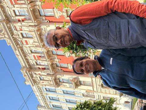       Smiling couple poses in front of an ornate red-and-cream Art Nouveau façade under a clear blue sky.
  