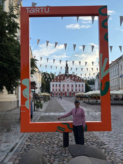       Traveler stands beneath a decorative orange frame on a cobblestone square facing Tartu’s pink town hall, with bunting strung overhead and cafés lining the plaza.
  