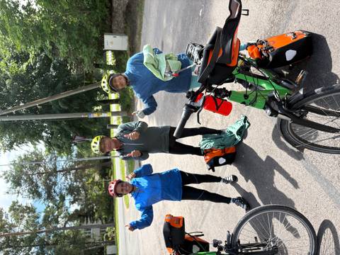       Three helmeted cyclists give thumbs-up beside their touring bikes in a sunny parking area framed by tall trees.
  