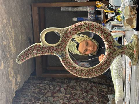       A craftswoman paints intricate designs on pottery, framed creatively through a decorated ewer.
  
