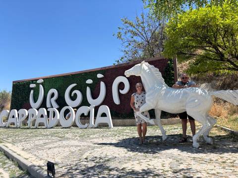       Two travelers pose beside a white horse statue by the Ürgüp Cappadocia sign on a sunny day.
  