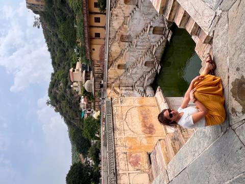       Smiling woman in white top and yellow skirt seated on stone ledge overlooking historic stepwell with green water.
  