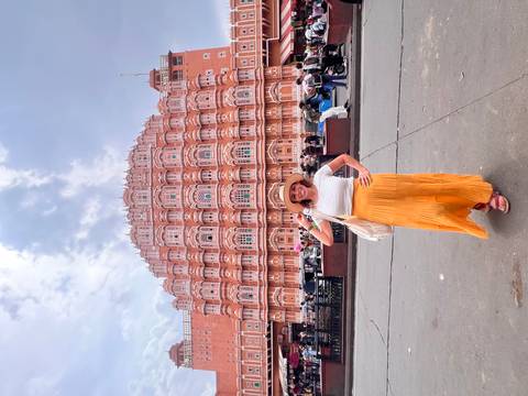       Traveler posing in front of ornate pink Hawa Mahal facade under dramatic cloudy sky.
  