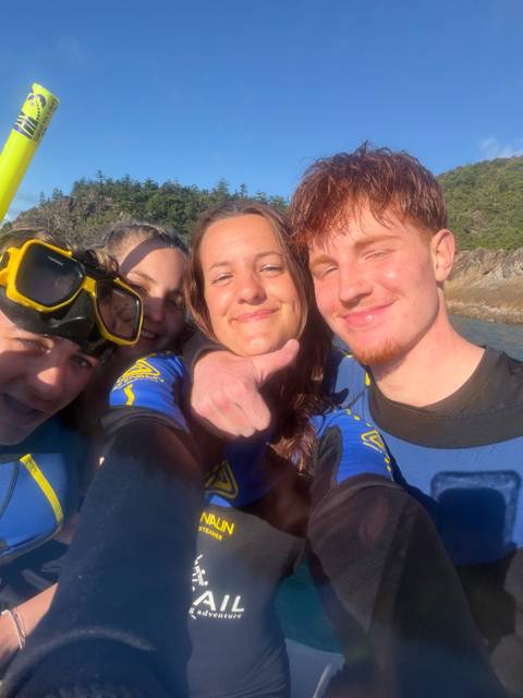       Close-up selfie of snorkellers in wetsuits with rocky shoreline behind
  