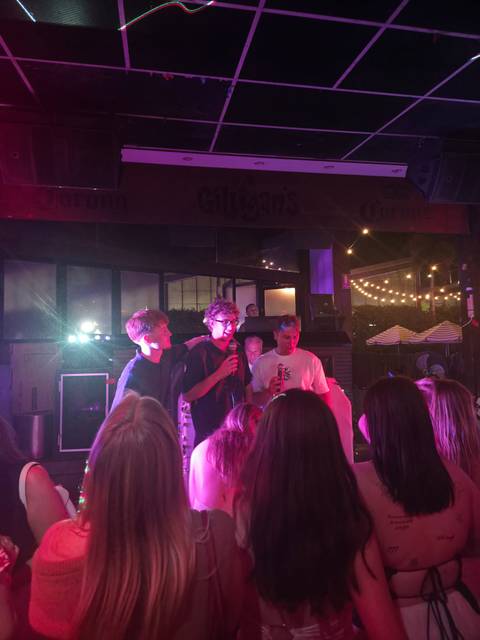       Young men singing on a small nightclub stage under purple and blue lighting while a crowd watches
  