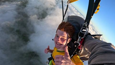       Smiling skydiver giving thumbs-up mid-air among white clouds with parachute straps visible
  