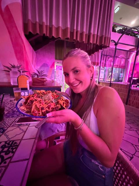       Smiling woman in a pink-lit restaurant holding a large plate of loaded nachos toward the camera
  
