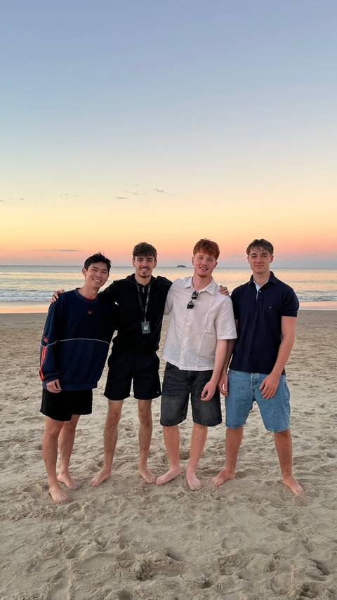       Four friends pose arm-in-arm on a beach at sunset with calm sea and orange sky behind them
  