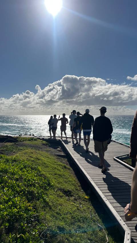       Group of people walking along a coastal boardwalk toward the bright ocean horizon under dramatic clouds
  