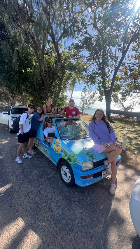       Smiling group gathered around a small open-top car painted with tropical graphics near the coast
  