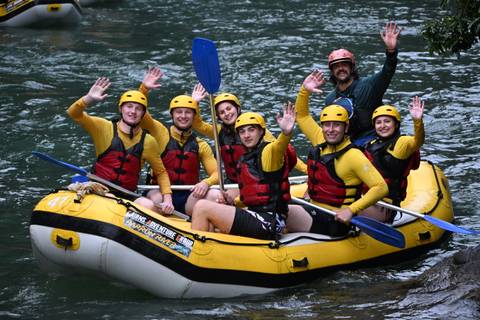       Raft full of paddlers in yellow helmets and red life jackets waving while floating on a green river
  