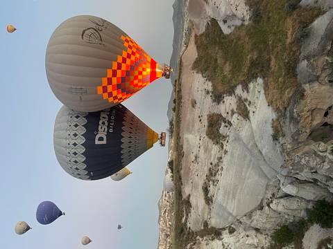       Two large balloons hovering low over Cappadocia’s white rock cliffs, one glowing with orange burner flame.
  
