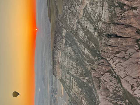       Orange sun peeks over Cappadocia’s distant horizon casting warm light on rugged cliff formations below.
  