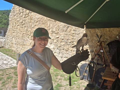       Smiling woman wearing a cap holds a falcon on a gloved hand beside a stone fortress wall and a small display stand.
  