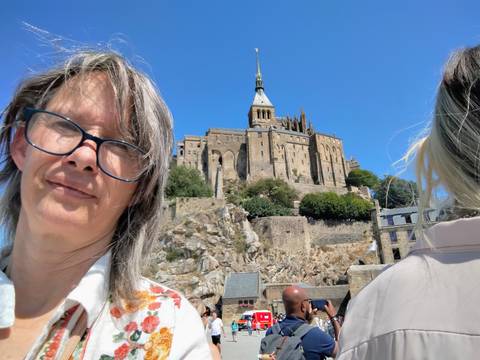       Selfie with a close-up tourist in front of Mont-Saint-Michel abbey rising against a blue sky.
  