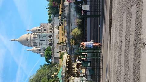       Woman poses at the base of Sacré-Cœur Basilica on Montmartre with carousel and gardens below.
  