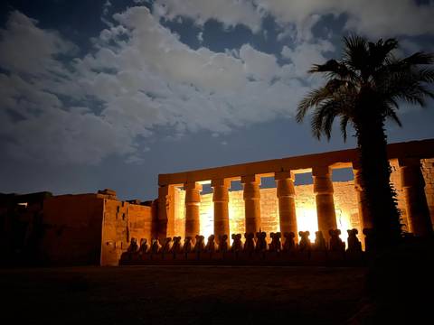       Night view of illuminated temple colonnade with dramatic lighting against a moonlit cloudy sky and palm silhouette.
  