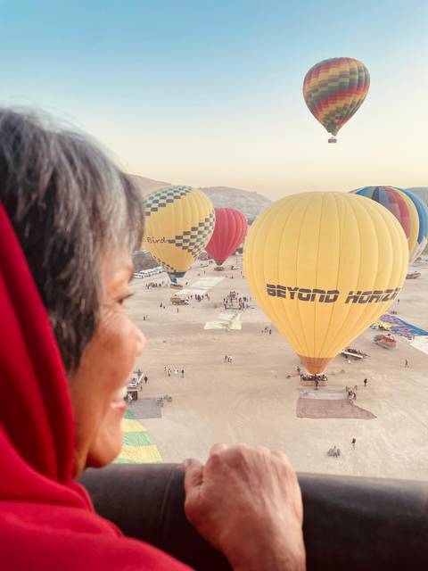       Hot air balloons rise over desert landscape at dawn with a passenger in red scarf watching from basket foreground.
  