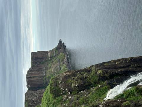       Dramatic sea cliffs plunging into calm grey water with overcast skies above.
  