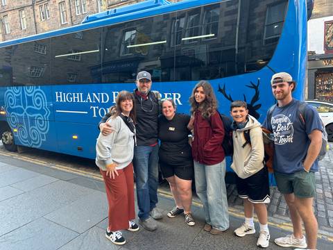       Smiling tour group posing in front of a blue Highland Explorer Tours coach on a city street.
  