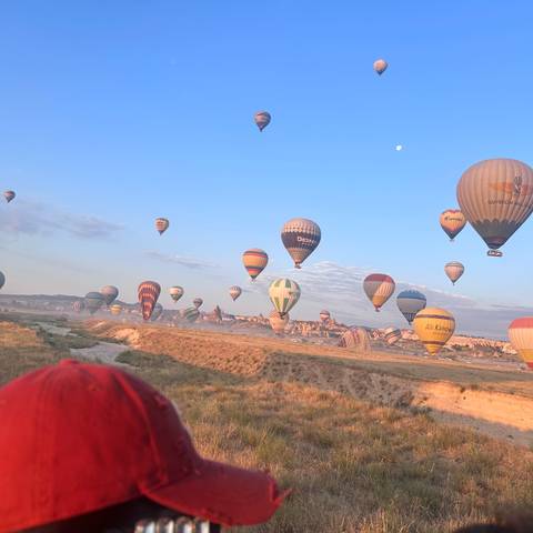       Morning sky filled with colourful hot-air balloons rising above Cappadocia’s dusty plains beneath a visible moon.
  