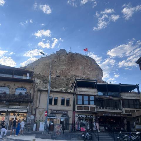       Rocky citadel topped by a Turkish flag rises above modern cafés and houses under blue skies.
  