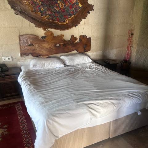       Simple double bed with rustic wooden headboard inside a stone-walled cave hotel room.
  