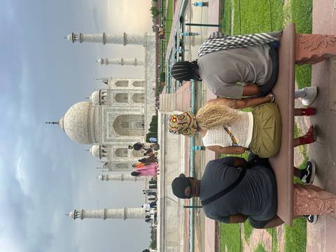       Three travellers sit on a bench admiring the iconic white marble Taj Mahal across its reflecting pools.
  