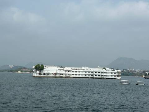       Lake Palace hotel floating on a calm lake with hazy mountains and city in the background.
  